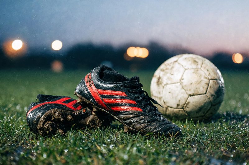 Abgetragene schwarz-rote Fussballschuhe und ein Ball auf nassem Rasen